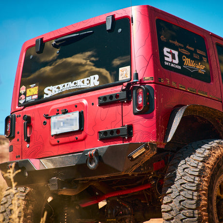Rear view of a red Jeep JK showcasing Tail Gunner Tail Lights installed on both sides. The lights are housed in black aluminum casings with red LED main lights and side markers, blending seamlessly with the vehicle’s design. The Jeep features a custom rear bumper, off-road modifications, and sponsor decals on the rear window. The image captures the vehicle in a rugged outdoor environment, emphasizing its off-road capability and style.