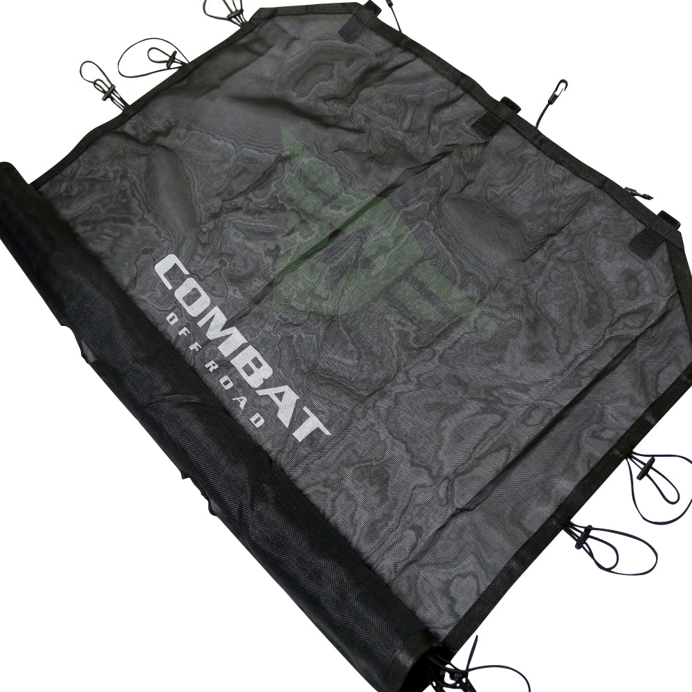 Angled view of a black mesh sunshade designed for a 4-door Jeep JK. The Combat Off Road logo is prominently displayed in white across the center of the fabric. The sunshade features durable construction with reinforced edges and multiple bungee cords with hooks around the perimeter, ensuring a secure fit to the vehicle's roll bars. The material provides shade while maintaining airflow for a comfortable interior.